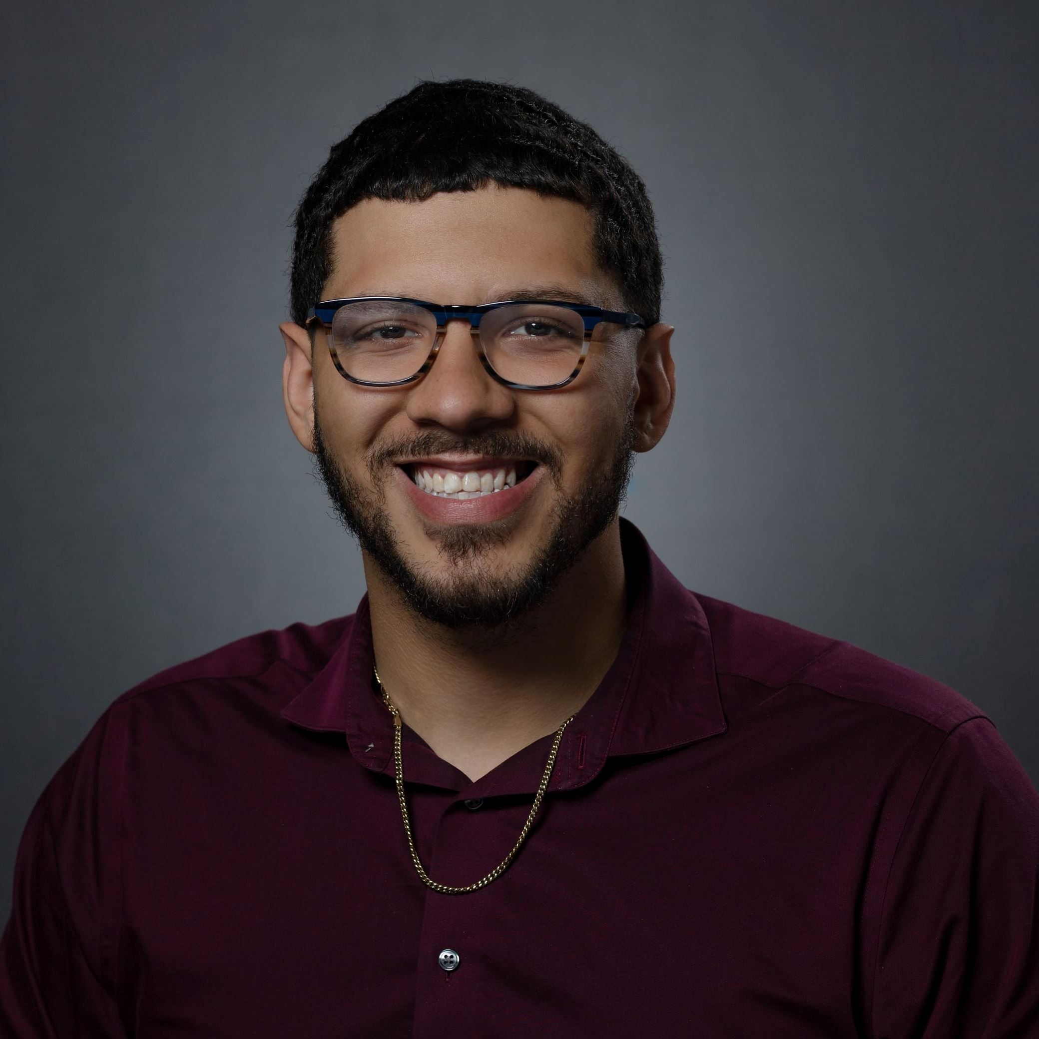 Headshot of Malik Frederick male with dark hair/beard and glasses wearing burgandy polo shirt