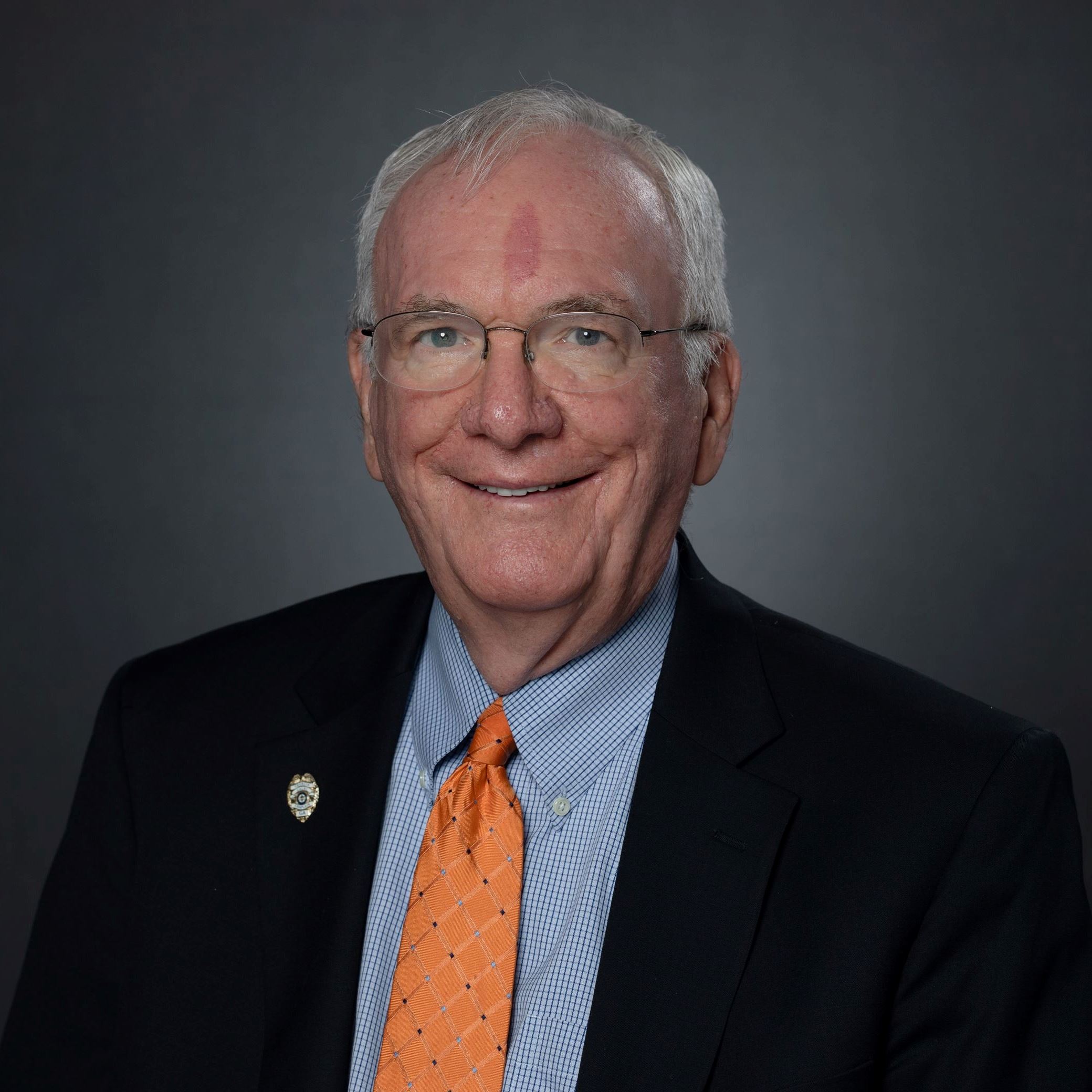 Headshot of Scott Batterton male with white hair and glasses in black suit orange tie